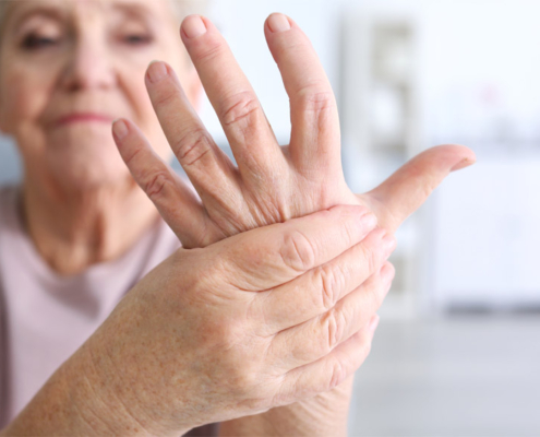 An elderly woman with arthritis grimacing while holding and massaging her left hand with her right hand.
