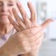 An elderly woman with arthritis grimacing while holding and massaging her left hand with her right hand.