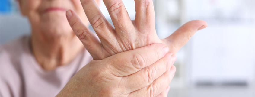 An elderly woman with arthritis grimacing while holding and massaging her left hand with her right hand.