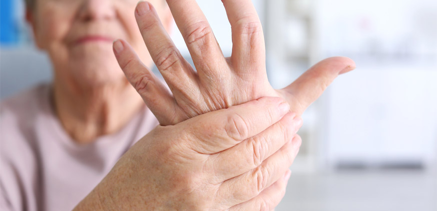 An elderly woman with arthritis grimacing while holding and massaging her left hand with her right hand.