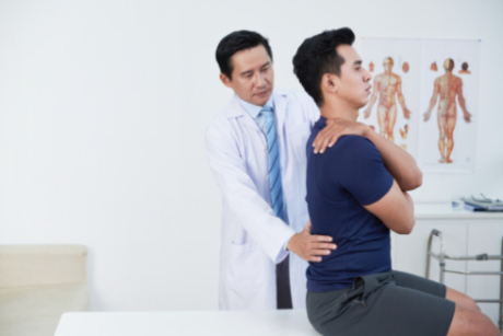 A male asian doctor examins a male asian patient's lower back as the patient sits up straight on an exam table.